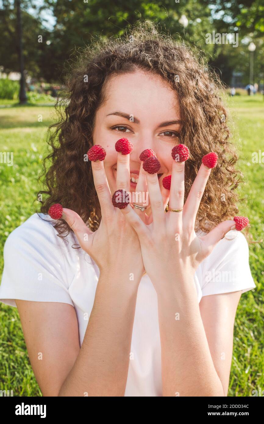 Verspielte junge Frau mit Himbeeren an den Fingern im Park während Sonniger Tag Stockfoto