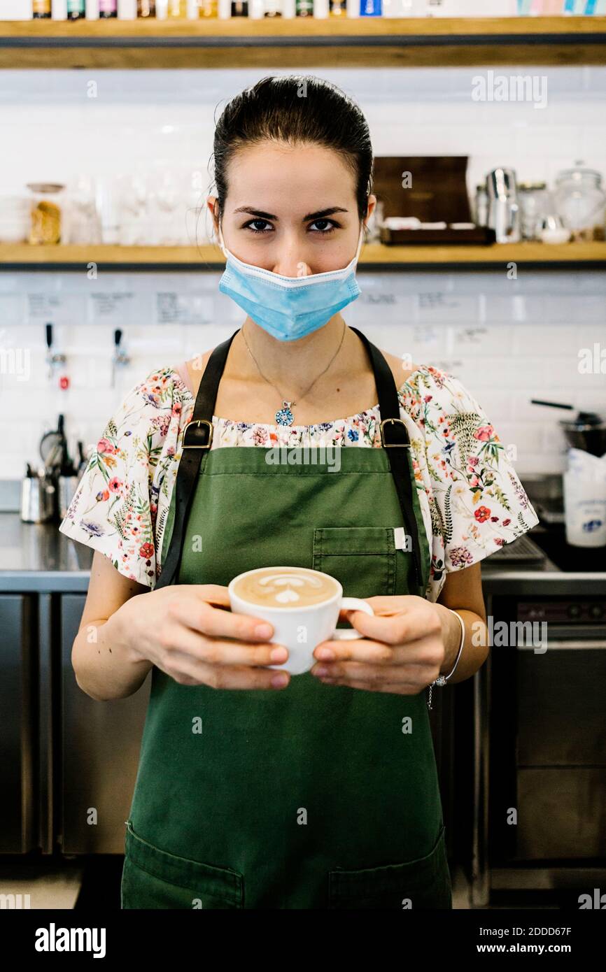 Junge Frau trägt Gesichtsmaske und Schürze mit Kaffeetasse Im Café stehen Stockfoto