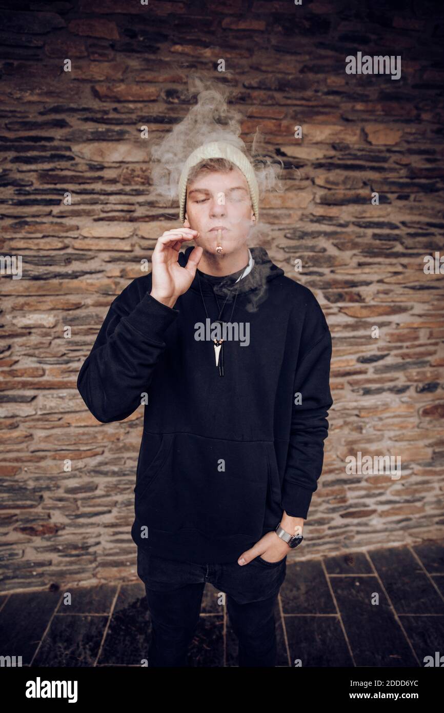 Junger Mann raucht mit Händen in Taschen, die gegen die Wand stehen Stockfoto