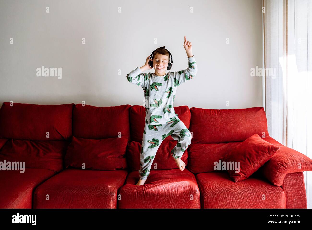 Fröhlicher Junge, der Musik über Kopfhörer hört, während er tanzt Sofa an der Wand Stockfoto
