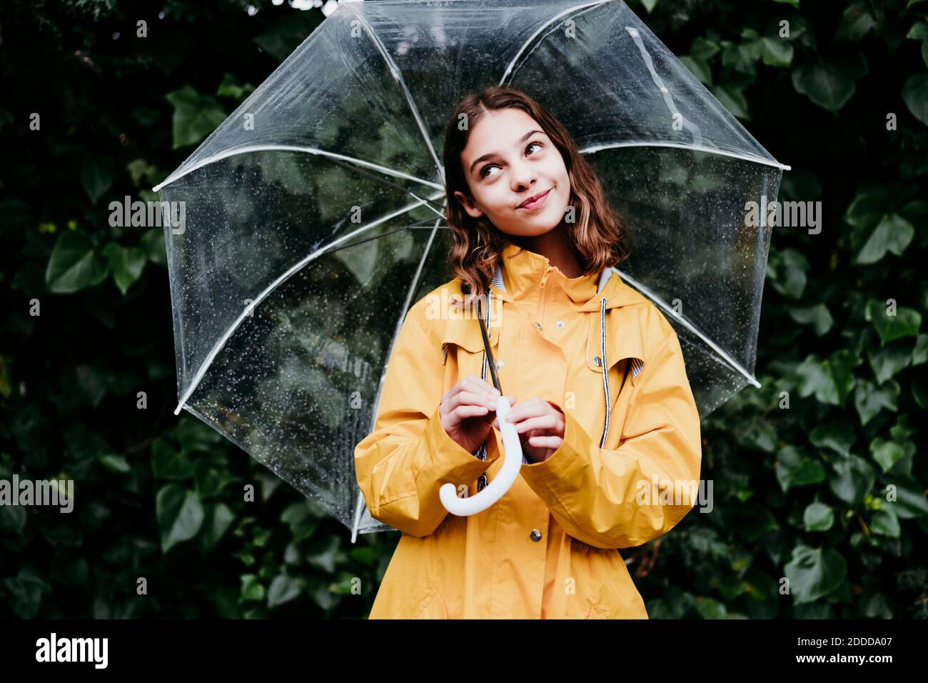 Nachdenkliches Mädchen lächelt, während Regenschirm gegen Blatt Wand hält Stockfoto