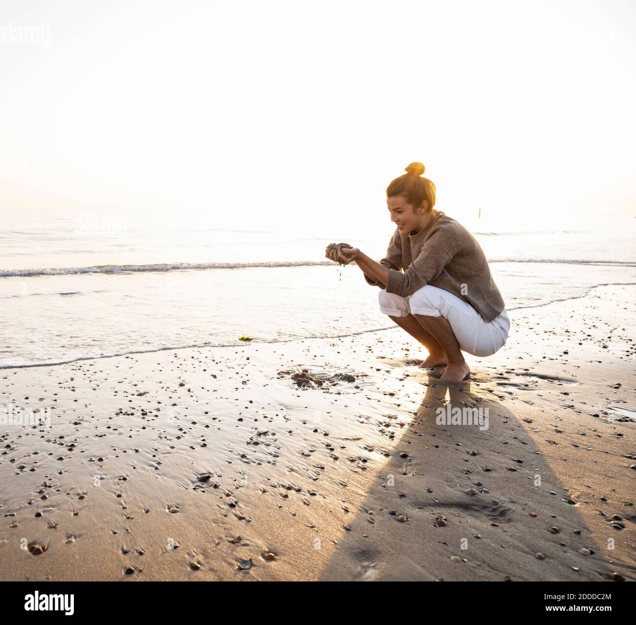 Schöne junge Frau hocken, während sie Sand am Ufer hält Strand bei Sonnenuntergang Stockfoto