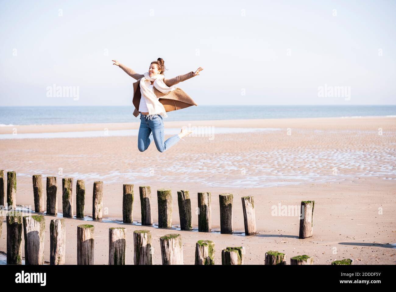 Glückliche junge Frau mit ausgestreckten Armen springen von Holzpfosten Am Strand an sonnigen Tagen Stockfoto