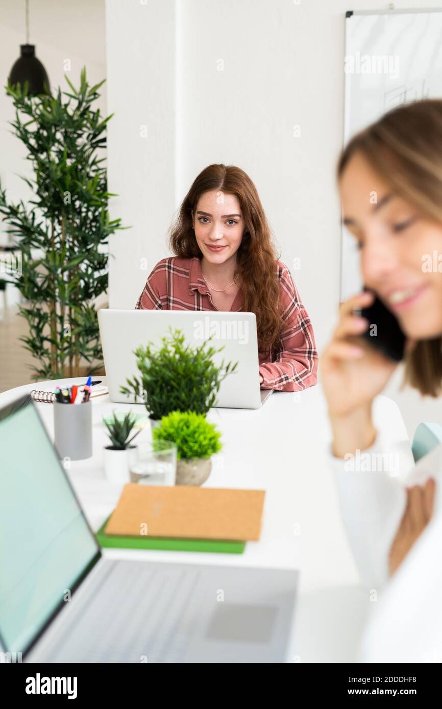 Lächelnde junge Frau, die am Laptop arbeitet und eine Geschäftsfrau spricht Mobiltelefon im Vordergrund im Büro Stockfoto