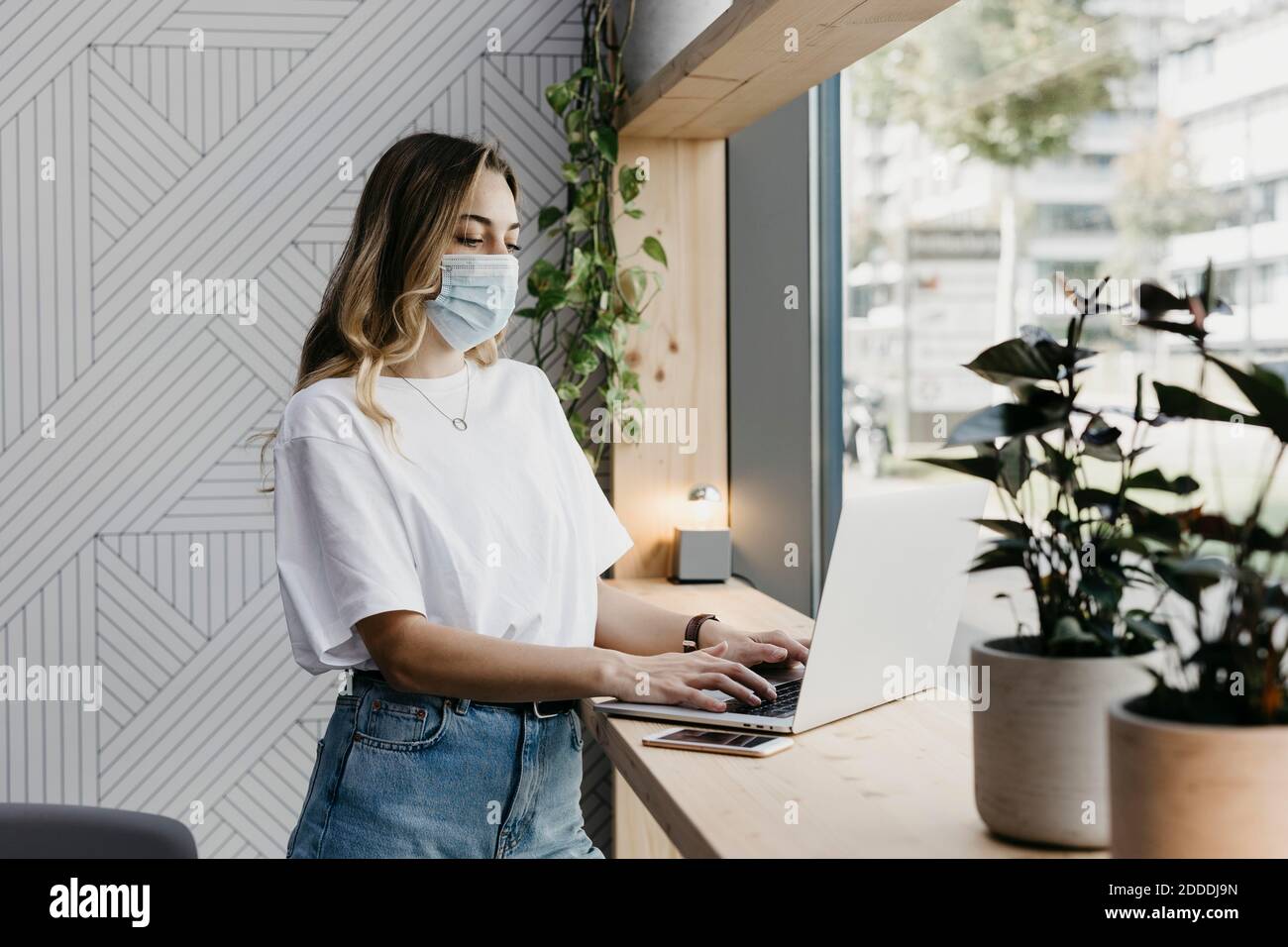 Frau trägt eine schützende Gesichtsmaske mit Laptop, während sie im Stehen ist Cafe während Covid-19 Stockfoto