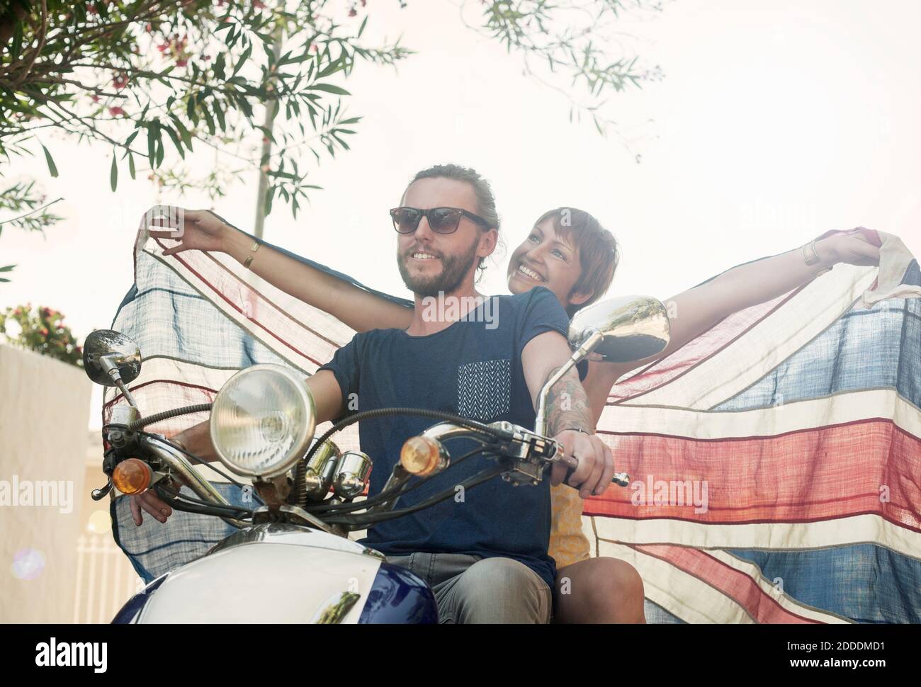 Lächelnde Frau mit ausgestreckten Armen und Nationalflagge im Sitzen Hinter Partner auf Motorrad im Sommer Stockfoto