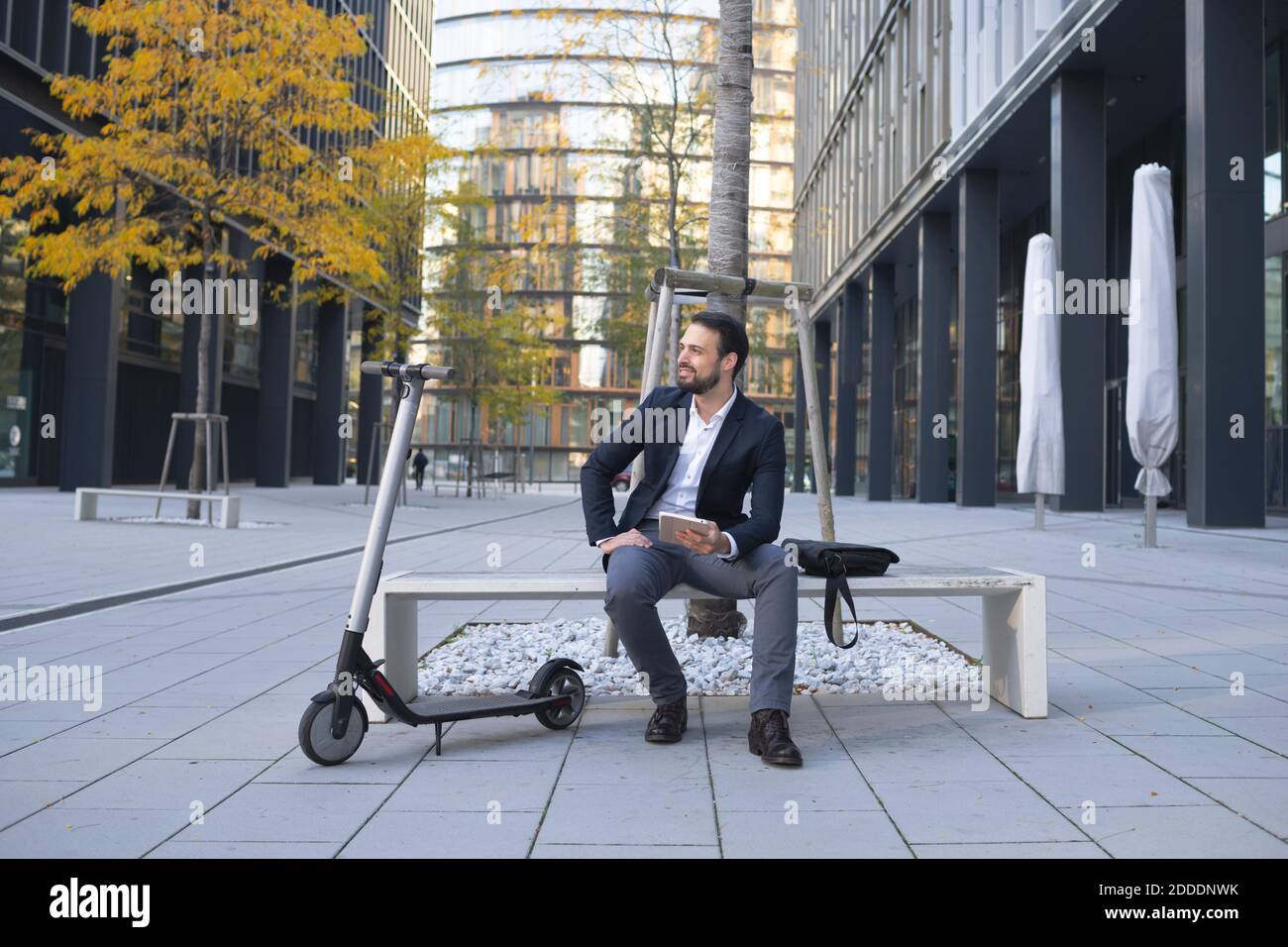 Unternehmer mit digitalen Tablet sitzen von E-Scooter auf Bank in Stadt Stockfoto