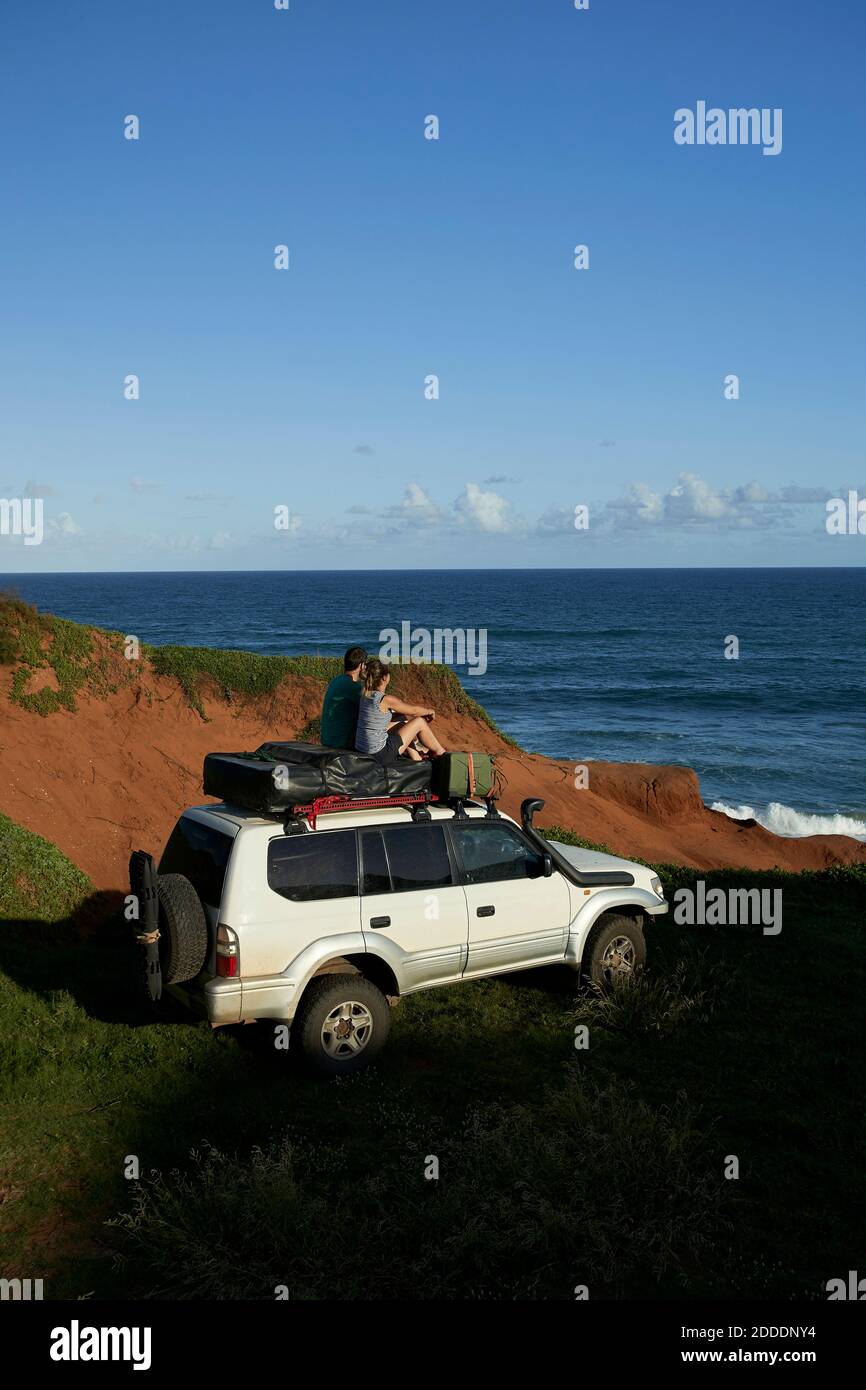 Mittelerwachsenes Paar, das auf dem 4x4-Dach sitzt, während es sich anschaut Meer gegen blauen Himmel Stockfoto