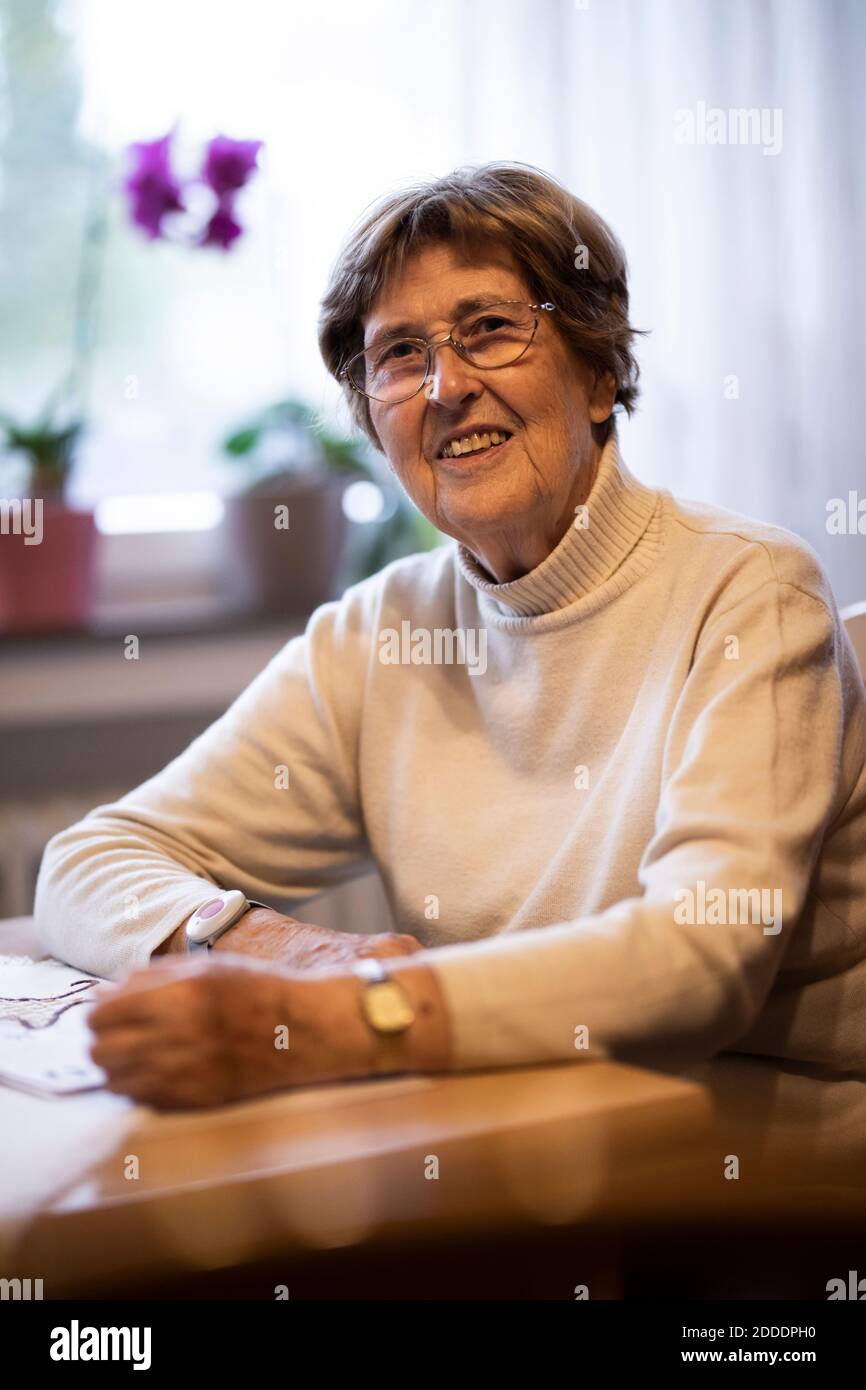 Lächelnd faltige Frau mit Nottaster am Handgelenk sitzend vorbei Tisch zu Hause Stockfoto