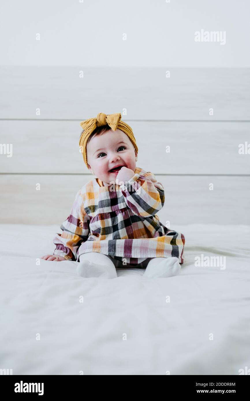 Lächelndes Baby mit dem Finger im Mund, der beim Sitzen wegschaut Auf dem Bett zu Hause Stockfoto