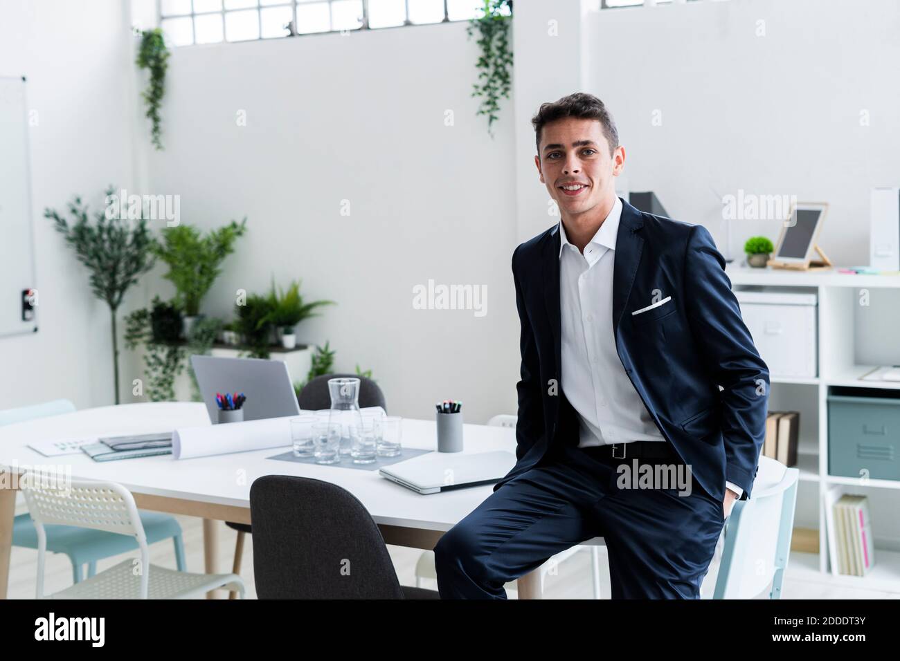 Lächelnder männlicher Unternehmer, der am kreativen Arbeitsplatz auf dem Schreibtisch sitzt Stockfoto