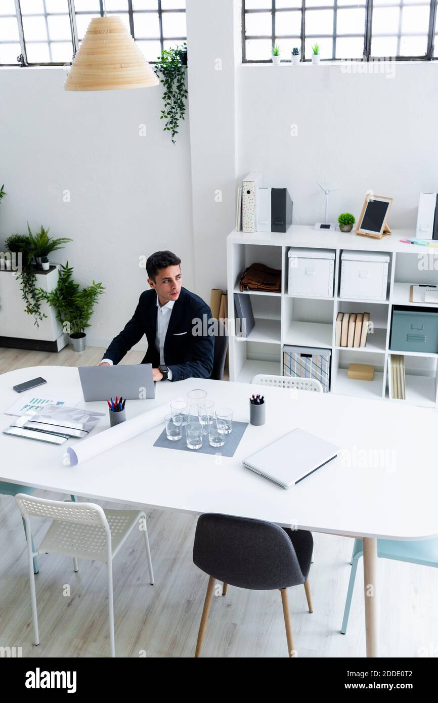 Nachdenklicher junger Unternehmer schaut weg, während er mit Laptop sitzt Am Schreibtisch am Arbeitsplatz Stockfoto