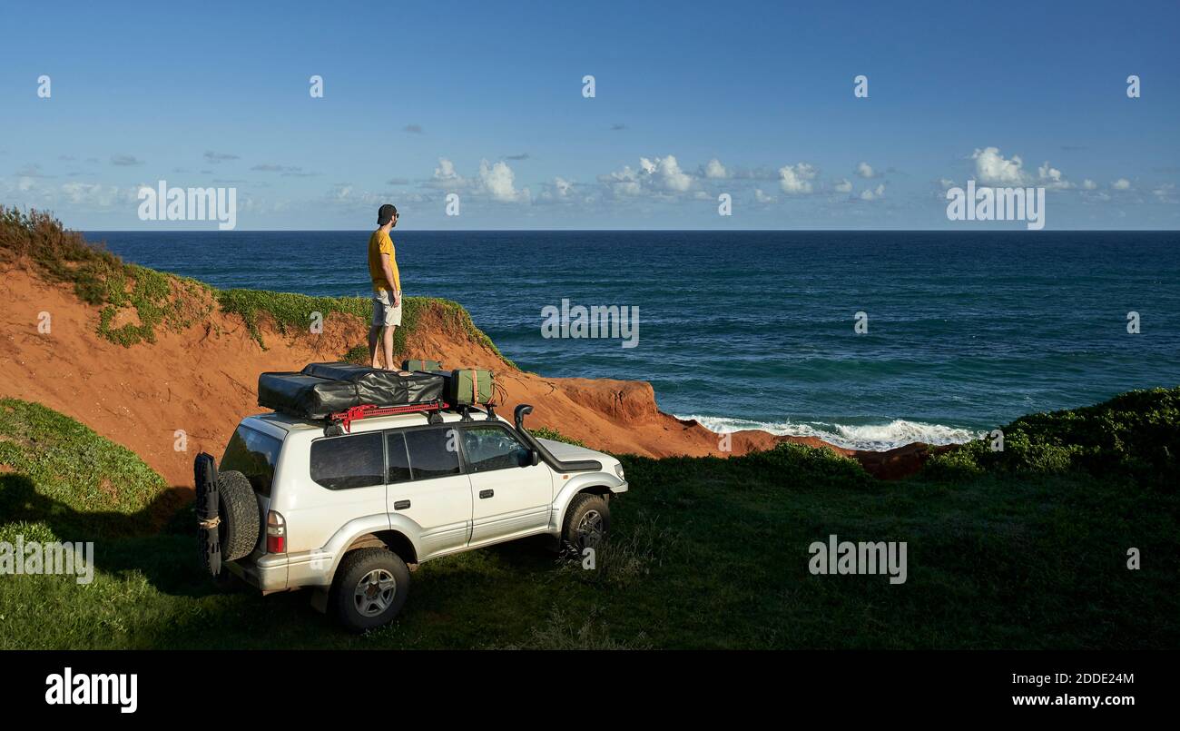 Männlicher Tourist, der auf dem 4x4-Dach steht und das Meer betrachtet Am blauen Himmel Stockfoto
