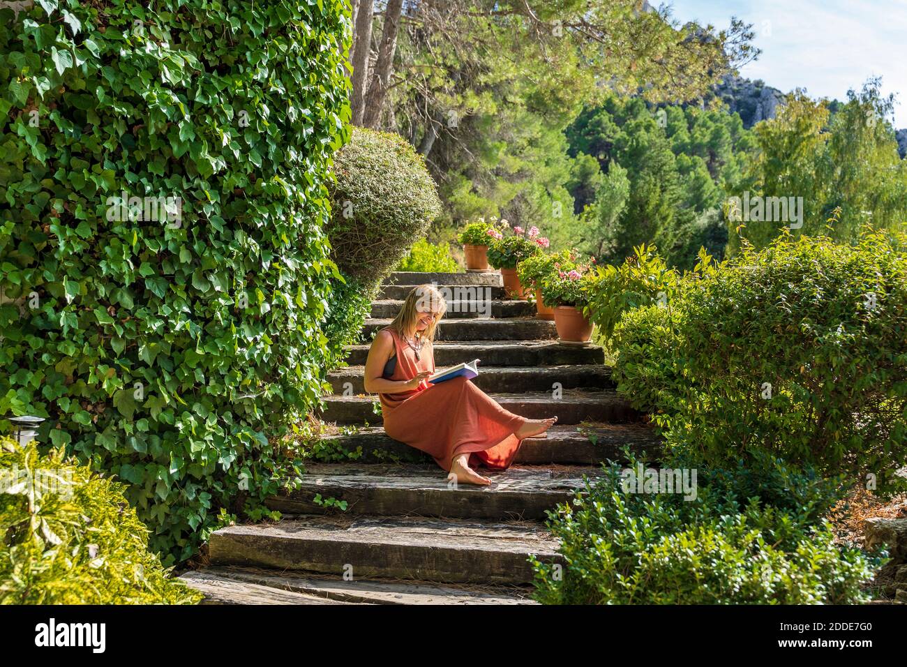 Reife Frau liest Buch, während sie auf Stufen inmitten von Grün sitzt Pflanzen bei Gesundheit Rückzug während sonnigen Tag Stockfoto