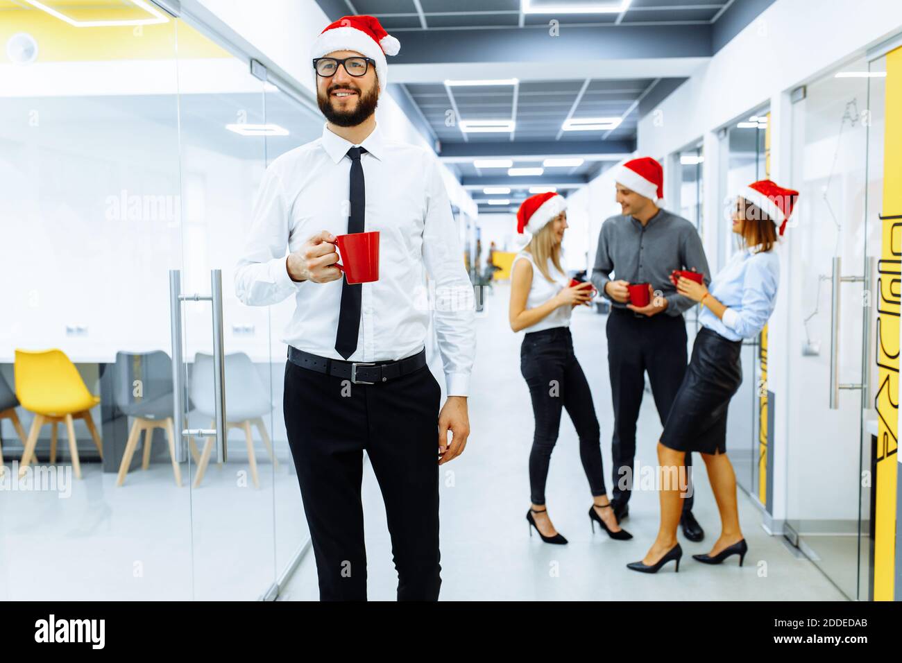 Junger Geschäftsmann trägt Weihnachtsmann Hut hält roten Becher mit Gruppe von Kollegen im Hintergrund während Kaffeepause im Büro. Neujahr und Chris Stockfoto