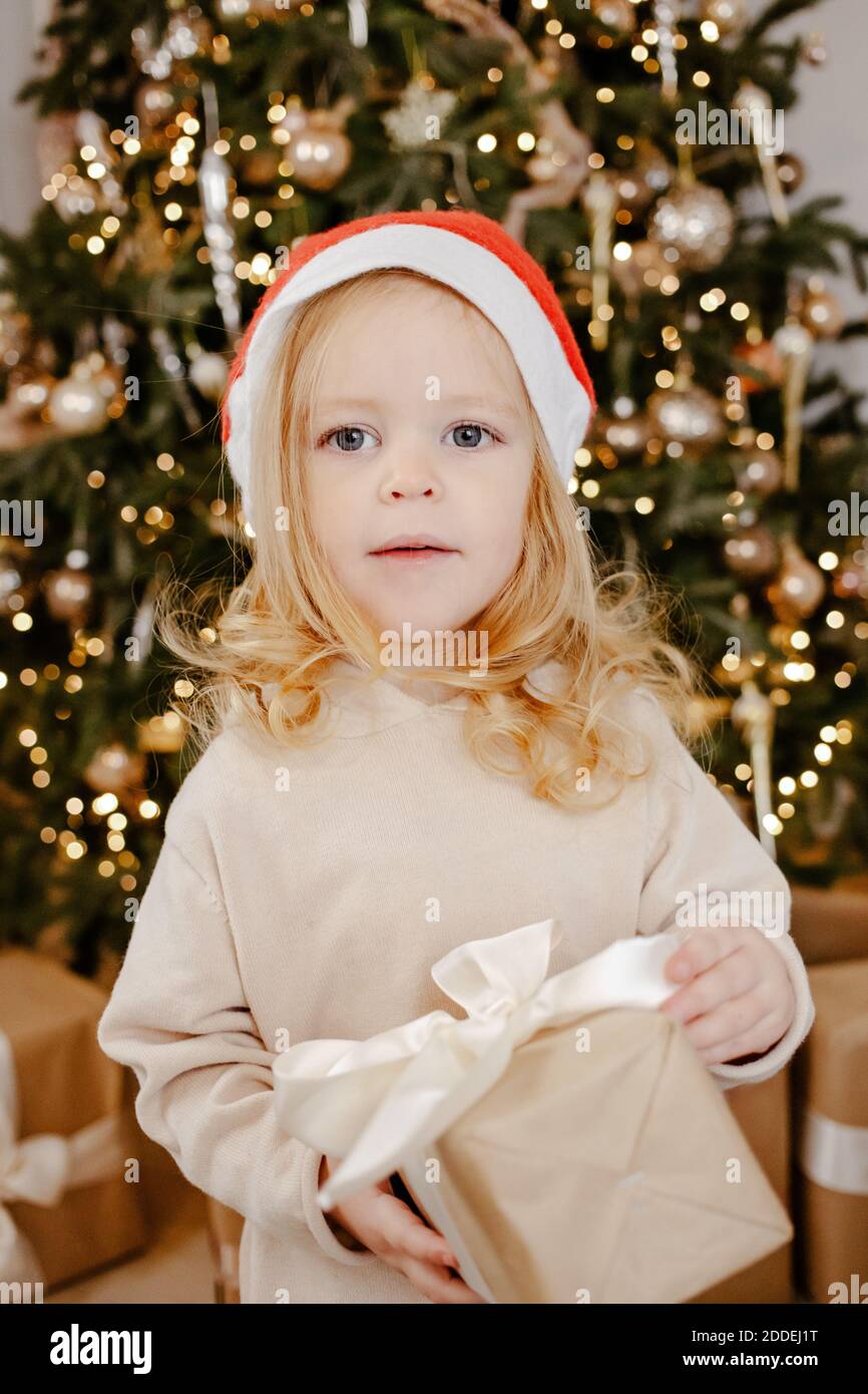 Nettes Baby Mädchen in Weihnachtsmann Hut unter Weihnachtsbaum mit Geschenkbox. Frohe Feiertage, Neujahr. Gemütlicher, warmer Winterabend zu Hause. Weihnachtszeit Stockfoto