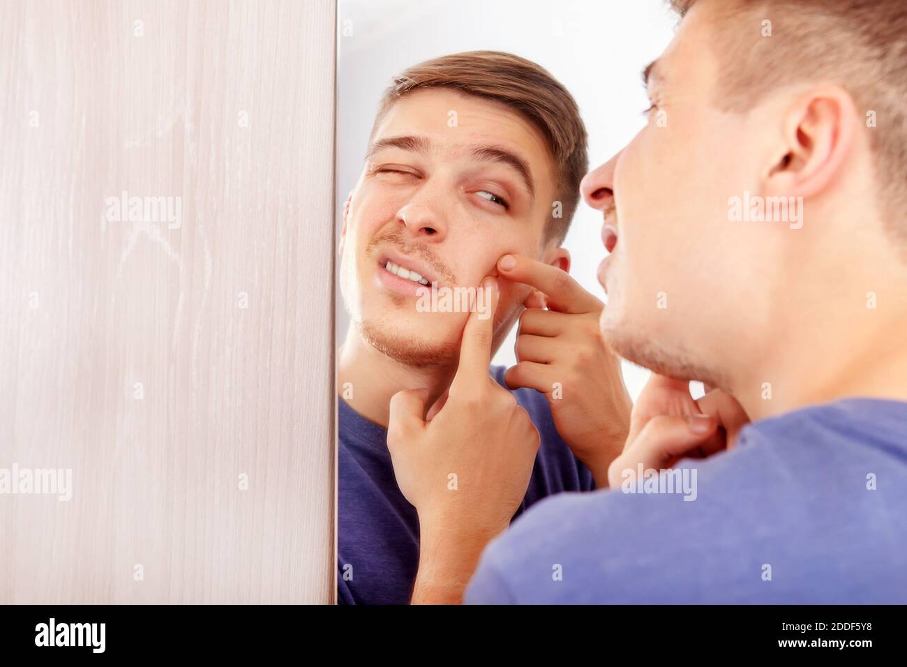 Junger Mann mit einem Pickel, der in der Reflexion sucht Spiegel im Zimmer Stockfoto