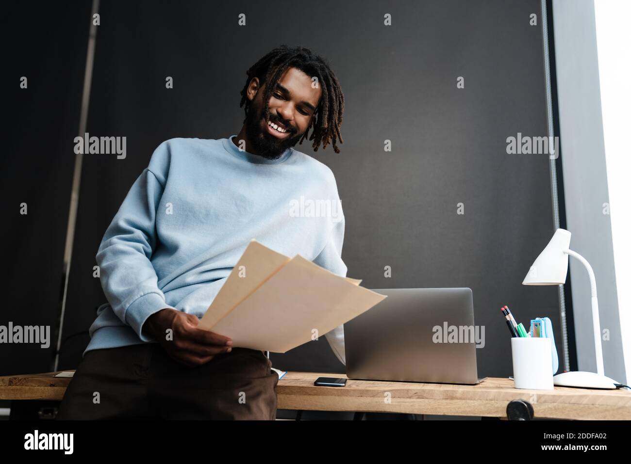 Lächelnder junger afrikanischer Mann, der Dokumente hält, während er sich auf ein stützt Schreibtisch Stockfoto