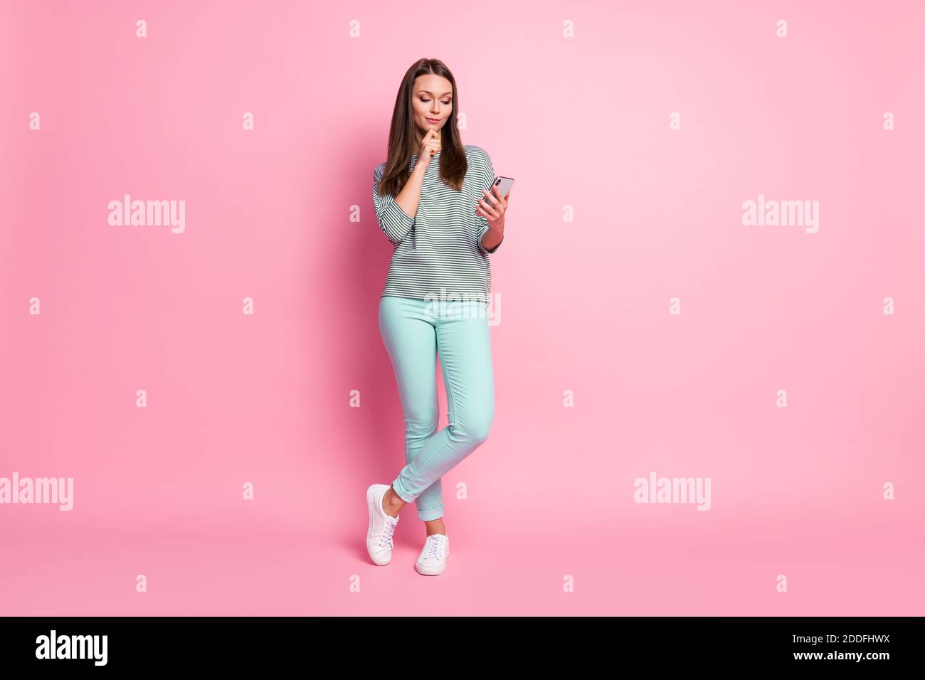 In voller Größe Foto von Interesse ziemlich braune Haare Frau stehen Look Telefon tragen blau Jeans Sneakers Pullover isoliert auf rosa Hintergrund Stockfoto