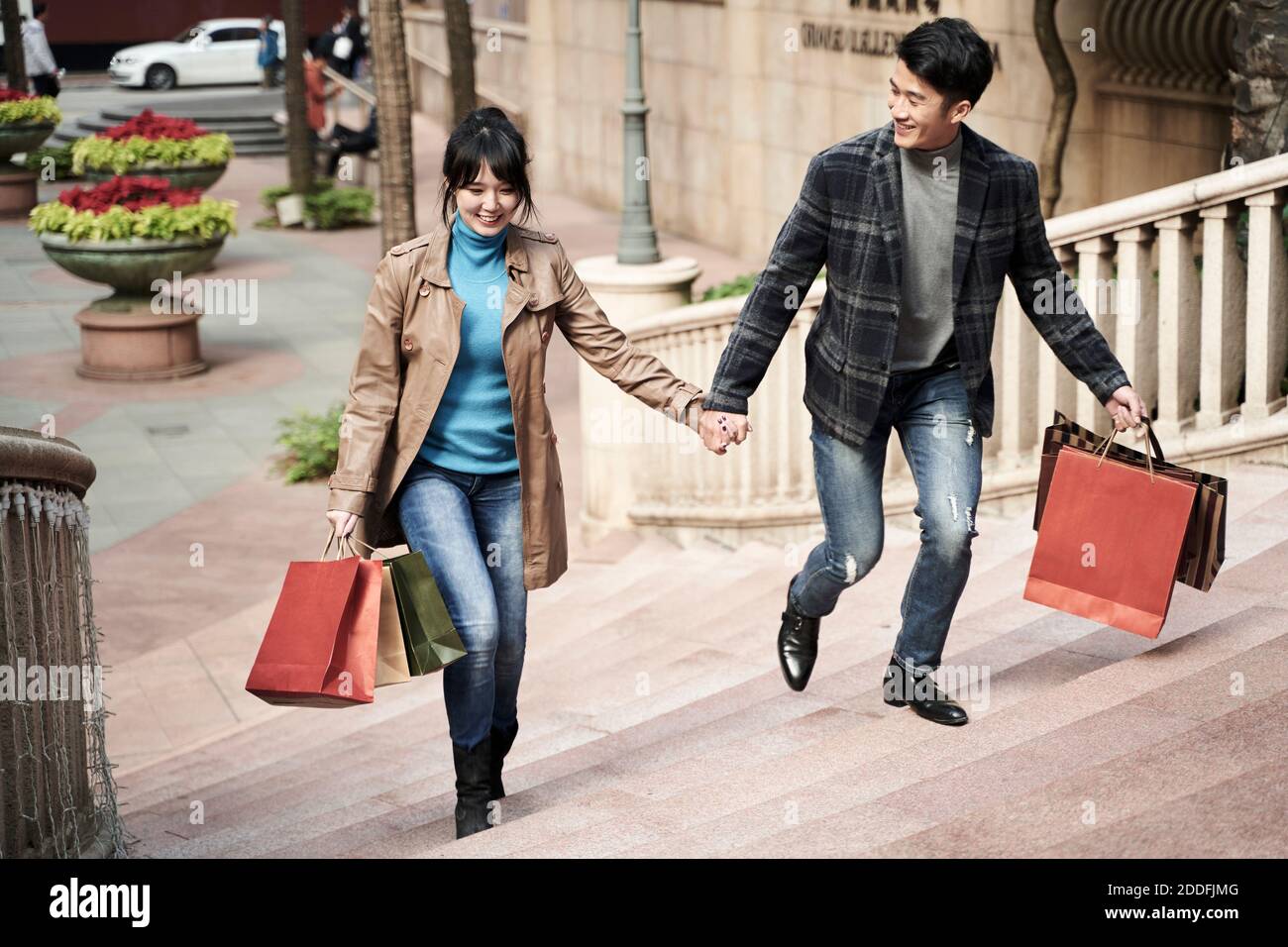 Junges asiatisches Paar mit Einkaufstaschen, die Hände halten, die auf Stufen gehen, glücklich und lächelnd Stockfoto