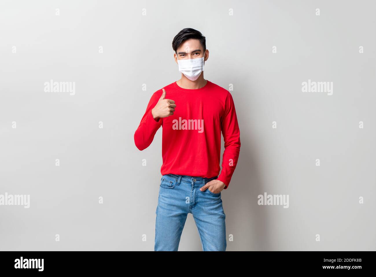 Junge asiatische trägt medizinische Maske und geben Daumen nach oben auf Hellgrauer Studiohintergrund Stockfoto