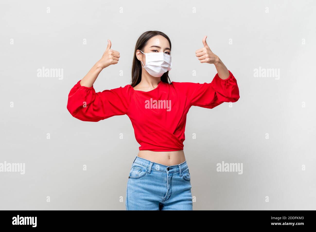 Junge asiatische Frau trägt Gesichtsmaske zu schützen COVID-19 und Daumen hoch auf hellgrauem Hintergrund isoliert Stockfoto