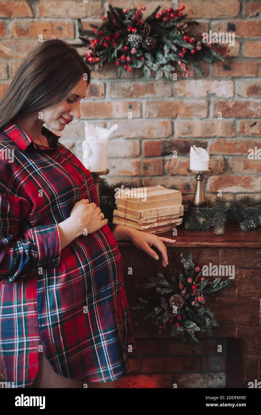 Schwangere Frau hält ihren Bauch neben einem gemütlichen Backstein Kamin in einem karierten roten Hemd. Gemütliche, rustikale Weihnachtsstimmung. Weihnachten, Winterschwangerschaft. Stockfoto