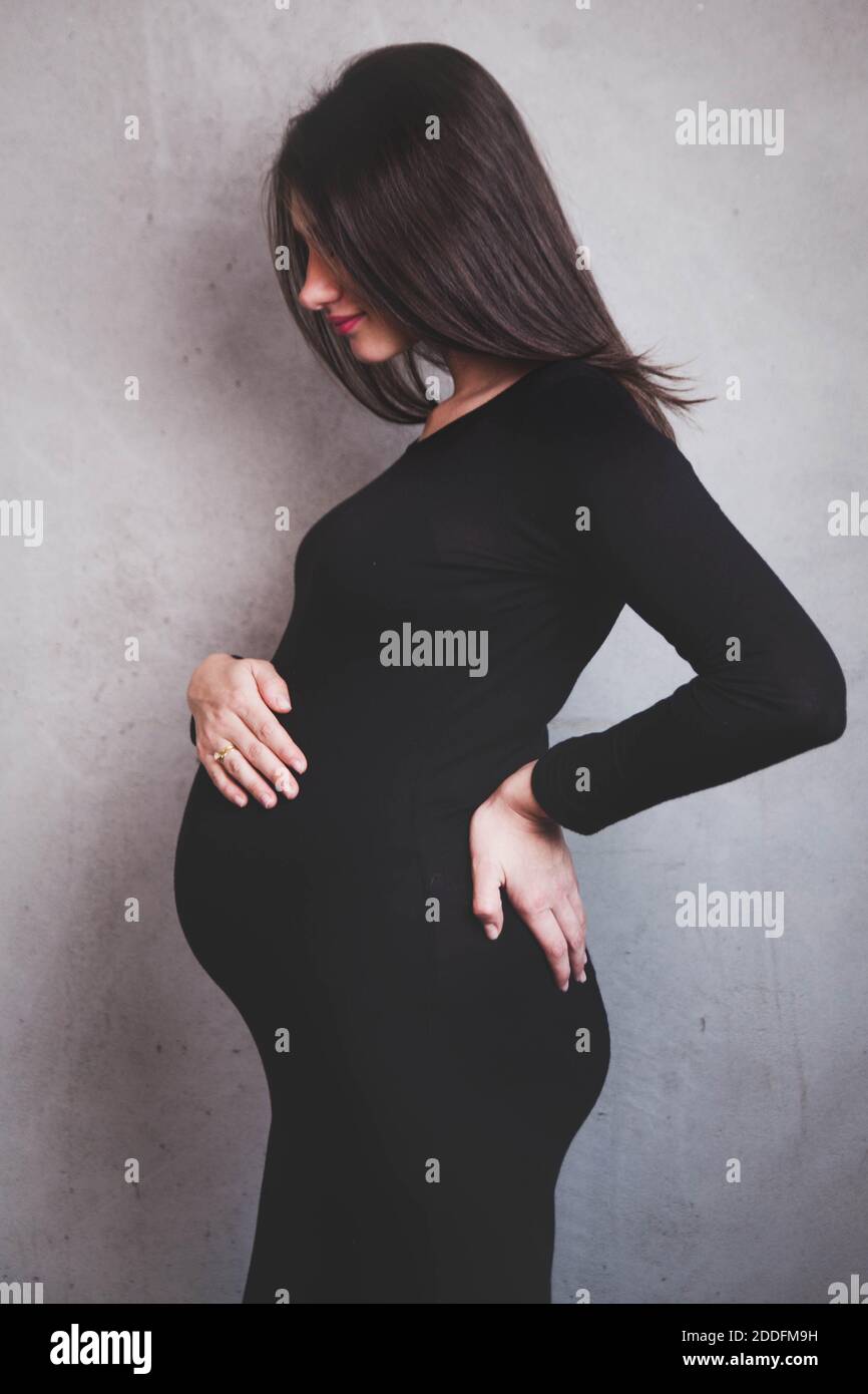 Porträt einer jungen dunkelhaarigen schwangeren Frau in einem schwarzen Kleid auf einem neutralen Hintergrund. Schwangerschaftsbump. Feminine erwartet Mutter Konzept. Stockfoto