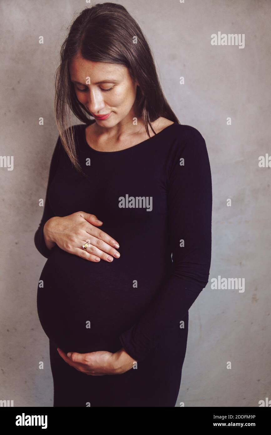 Porträt einer jungen dunkelhaarigen schwangeren Frau in einem schwarzen Kleid auf einem neutralen Hintergrund. Schwangerschaftsbump. Feminine erwartet Mutter Konzept. Stockfoto