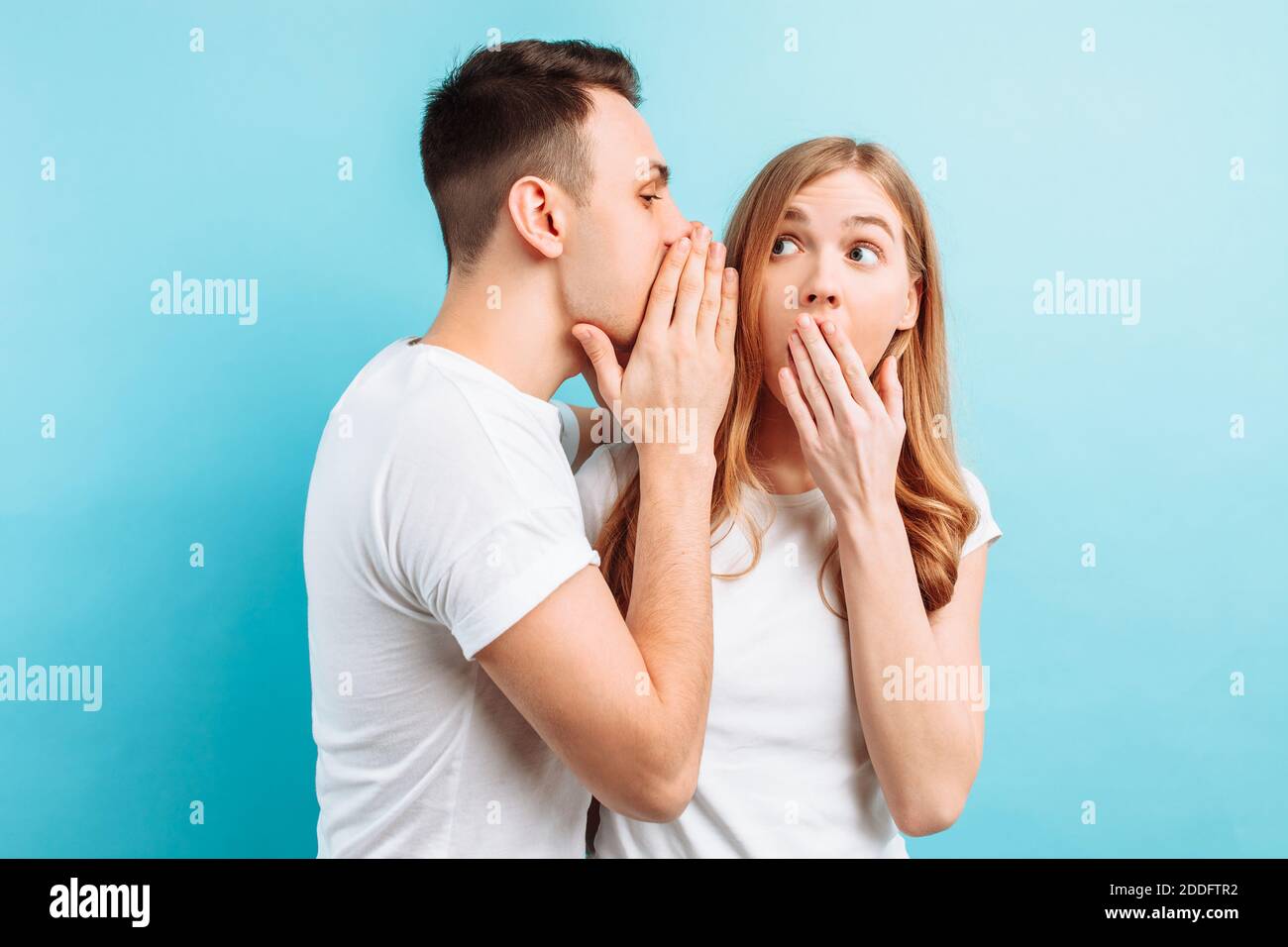 Ein Mann flüstert einer Frau ins Ohr und erzählt ihr etwas Geheimes, schockierend, auf blauem Hintergrund. Stockfoto