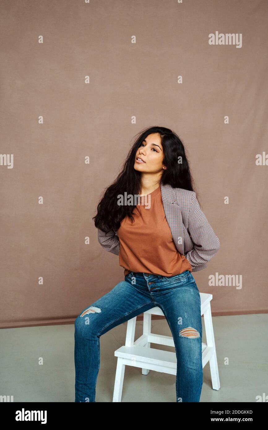 Stilvolle ethnische weibliche Modell in trendigen smart casual Outfit sitzen Auf dem Stuhl und wegschauen Stockfoto