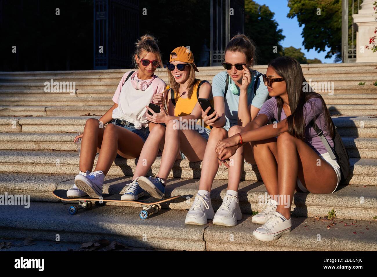 Fröhliche junge trendige multirassische Freundinnen in Sonnenbrillen Surfen Handys Während Sie sich an sonnigen Sommertagen auf Steintreppen ausruhen In der Stadt Stockfoto