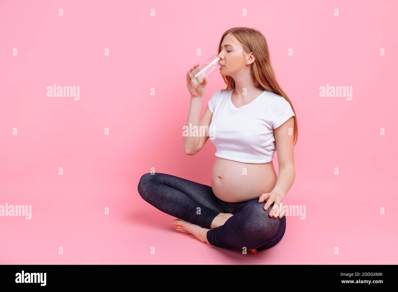 Schwangere Mädchen hält ein Glas Wasser auf dem Hintergrund eines schwangeren Bauch, auf einem rosa Hintergrund, das Konzept der Menge an Wasser verbraucht Stockfoto