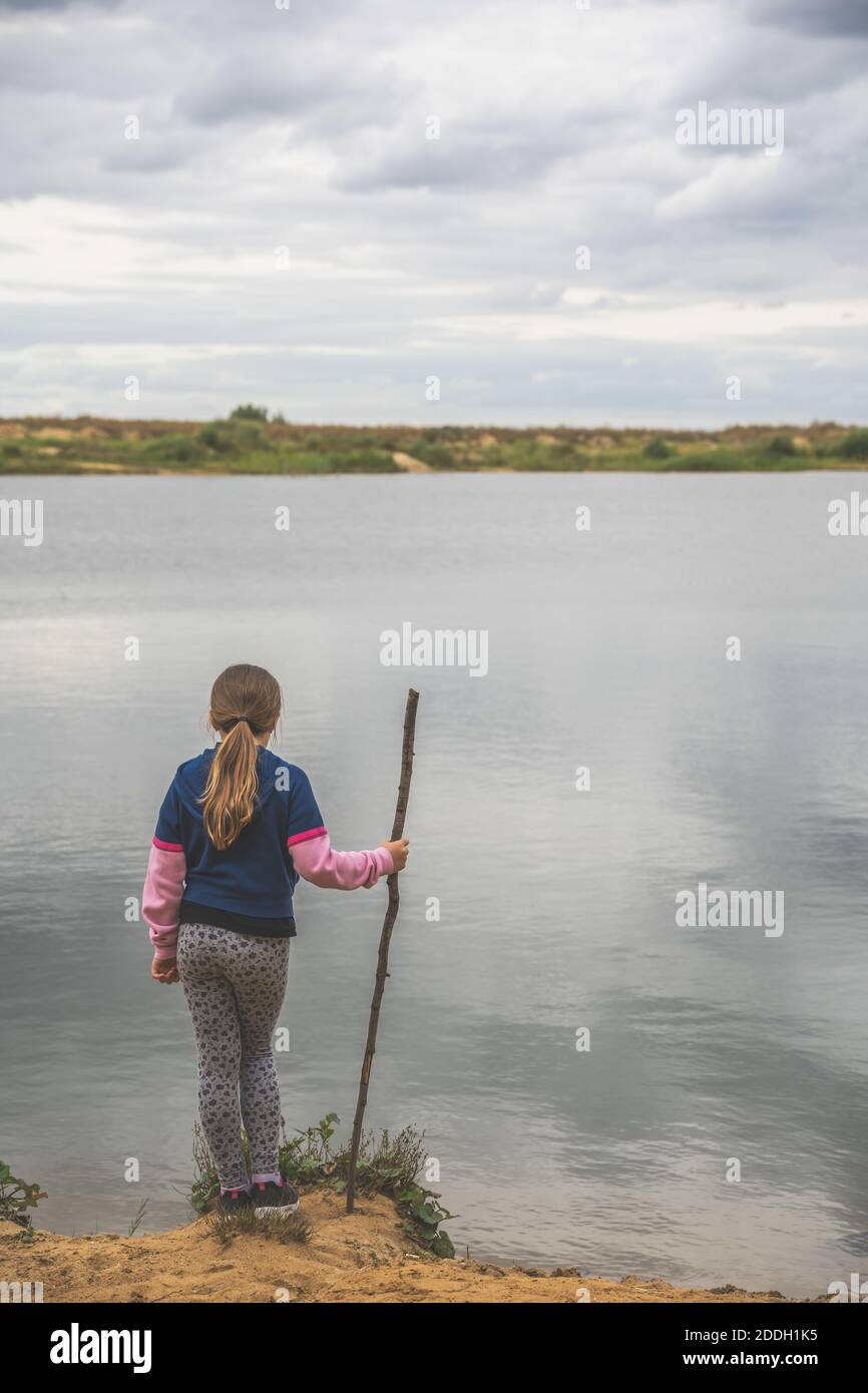 Ein flacher Fokus eines Kindes mit einem Holzstab Zum Angeln stehen in der Nähe des Sees unter einem bewölkten Himmel Stockfoto