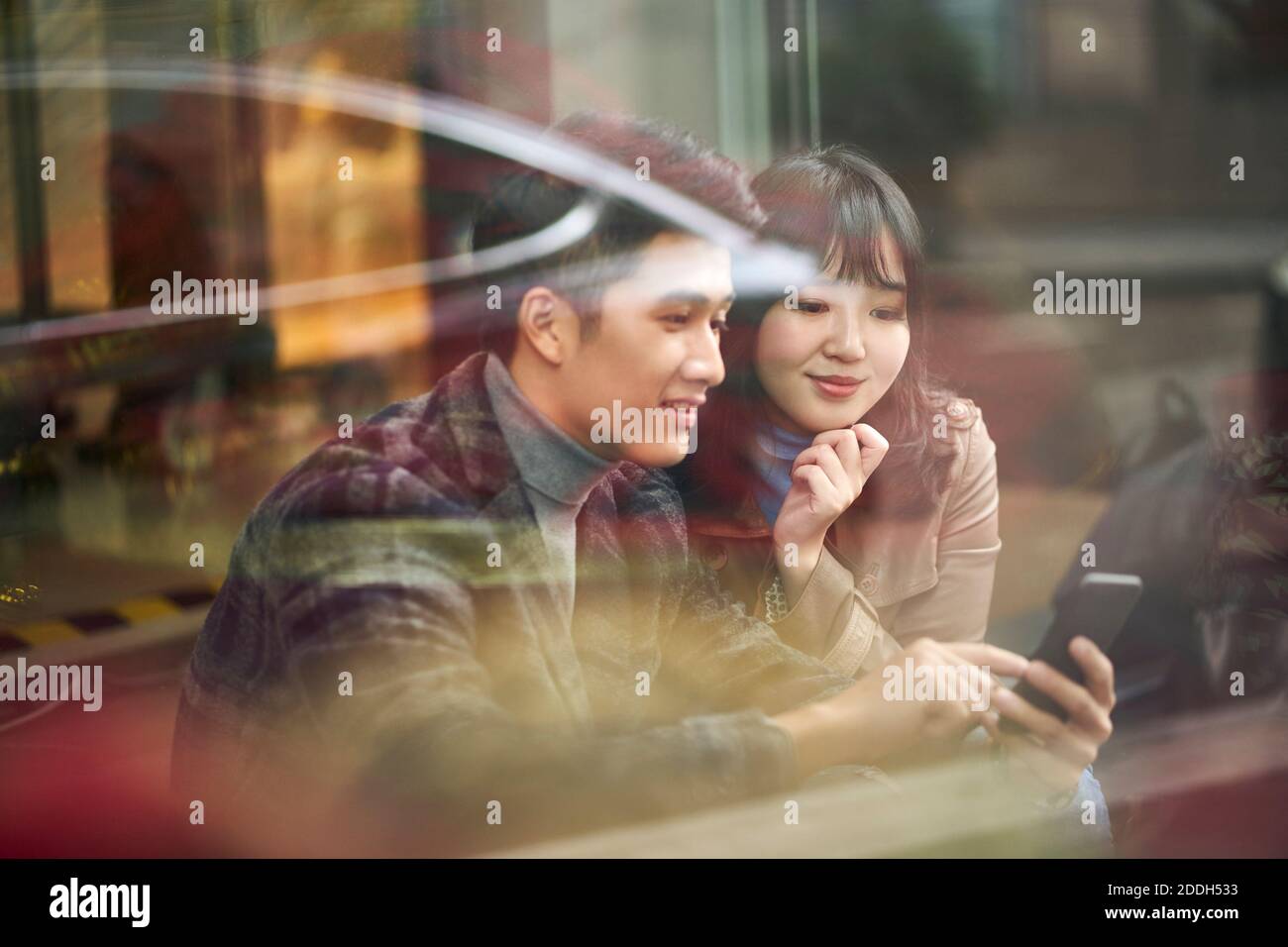 Durch die Glasaufnahme eines jungen asiatischen Paares sitzend Auf dem Boden Blick auf Handy Stockfoto