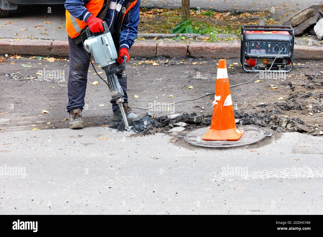 Ein Straßenarbeiter in reflektierender Kleidung zerschlägt alten Asphalt mit einem elektrischen Presshammer in der Nähe eines Schachtelschlochs. Stockfoto