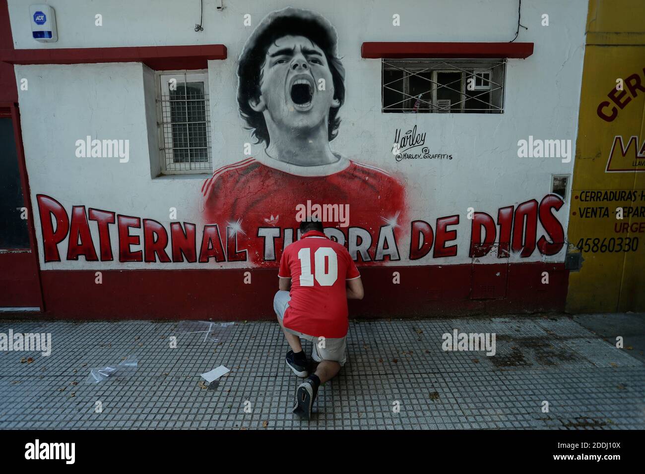 Buenos Aires, Argentinien. November 2020. Ein Fan, der ein Trikot des Argentinos Juniors aus dem Jahr 10 trägt, kniet mit Blumen vor einem Wandbild, das Diego Mardona darstellt, der im Alter von 60 Jahren starb. Der dribbelnde Künstler wuchs in ärmlichen Verhältnissen am Stadtrand von Buenos Aires auf und wurde schon in jungen Jahren vom First Division Club Argentinos Juniors entdeckt. Quelle: Gustavo Ortiz/dpa/Alamy Live News Stockfoto