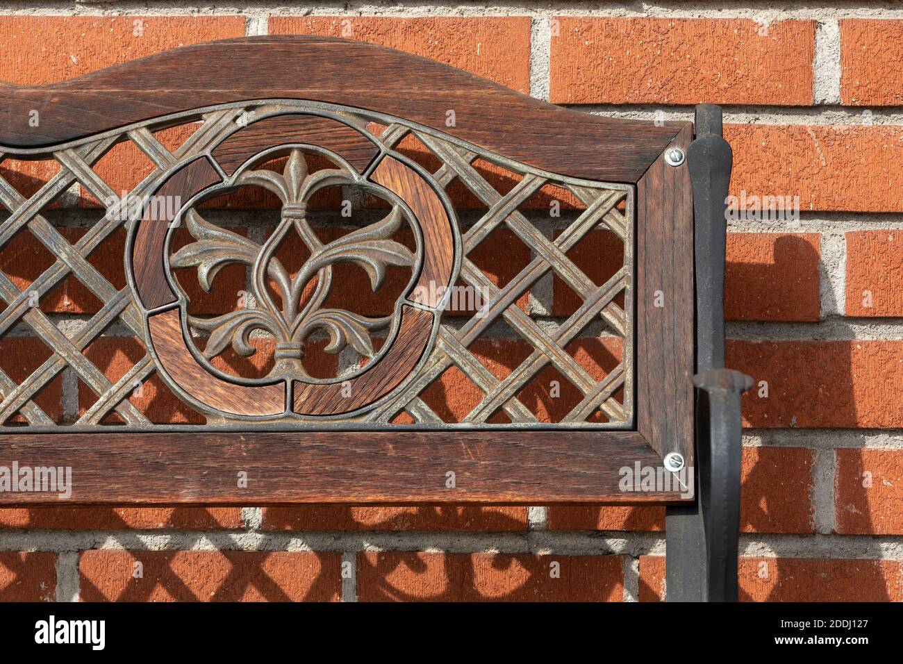 Nahaufnahme einer verzierten, verwitterten Holz- und Eisenbank mit eingelegtem Fleur de Lis-Muster, die einen starken Schatten an eine orangefarbene Ziegelwand wirft. Stockfoto