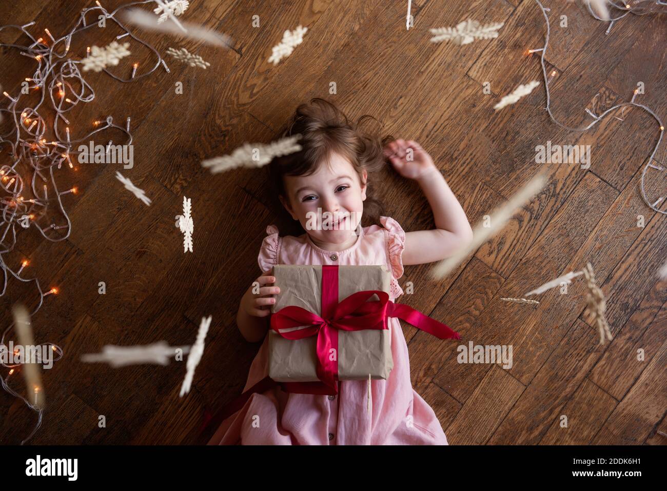 Kleines glückliches Mädchen in einem rosa Kleid liegt auf dem Holzboden. Hält eine Weihnachtsbox in Bastelverpackung, ein Weihnachtsgeschenk mit einem roten Band. Schneeflocken h Stockfoto