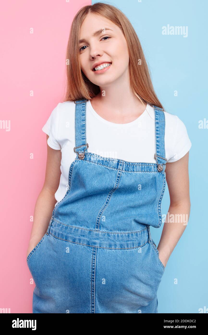 Eine schöne junge schwangere emotionale Frau in einem Jeansjumpsuit Posen auf einem isolierten rosa und blauen Hintergrund Stockfoto