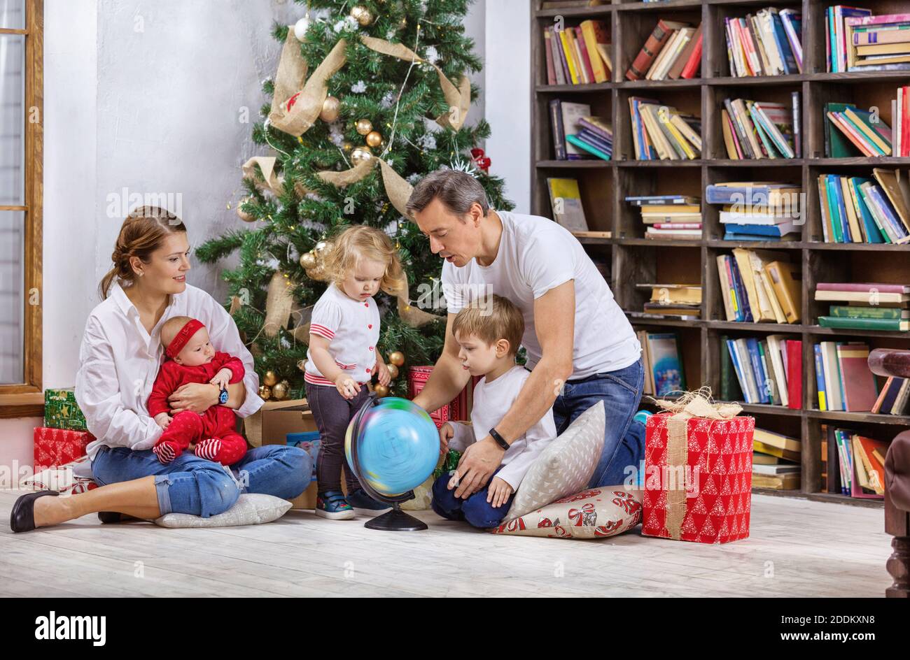 Familie mit drei Kindern neben Weihnachtsbaum zu Hause. Kinder und Vater untersucht Globus, Mutter hält Baby Tochter. Stockfoto