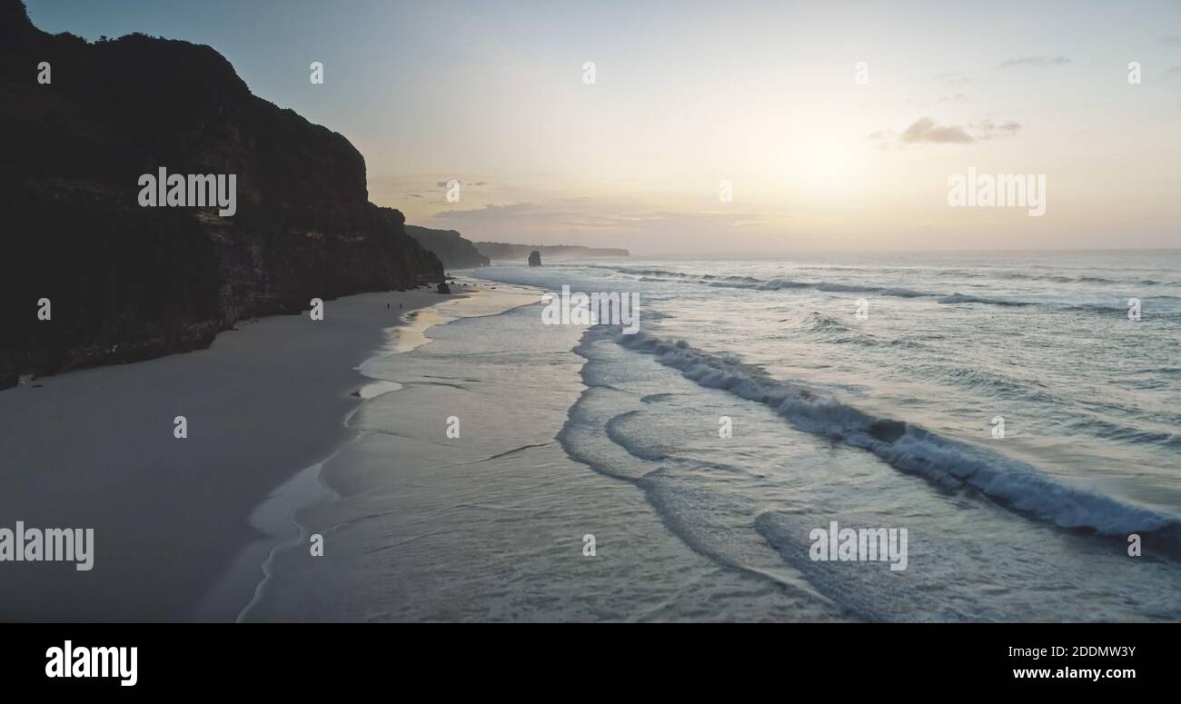 Sonnenaufgang am welligen Meer Klippe Küste Luftbild. Sonnenaufgang am dunklen Felsen, Meeresbucht Wasser gewaschen Sandstrand von Bawana, Sumba Insel, Indonesien. Entspannen Sie sich bei sanftem Licht in der ruhigen Meereslandschaft. Filmische Drohne erschossen Stockfoto