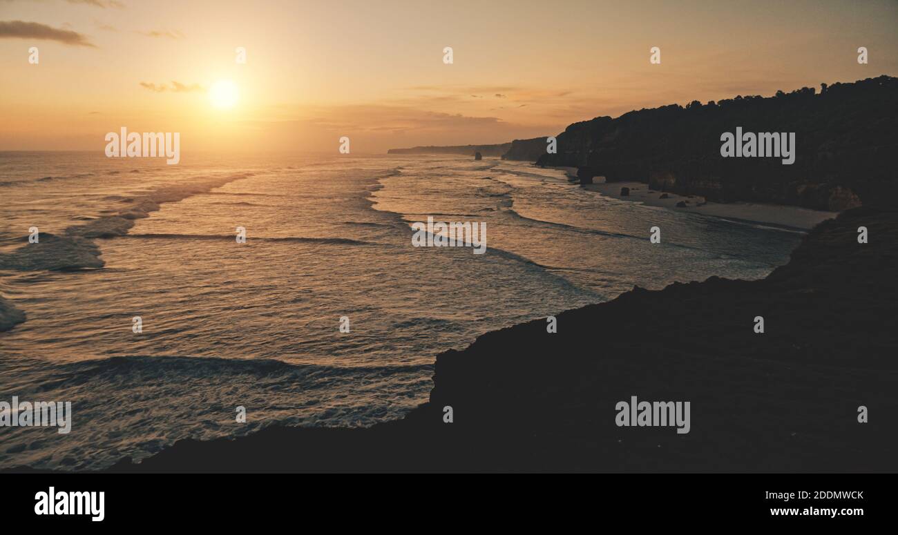 Entspannen Sie sich ruhige Seestück Sonnenuntergang Silhouette des Ozeans Klippe Küste Luftbild. Die kinoreife Sonne erhellte sich an der dunklen Felswand. Erstaunliche Meeresbucht Wellen Wasser gewaschen Sandstrand von Bawana, Sumba Insel, Indonesien Stockfoto