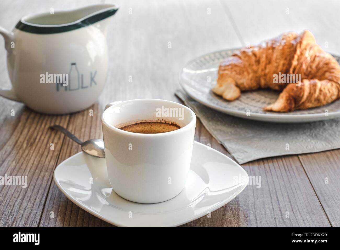 Horizontale Ansicht einer weißen Tasse Espresso mit Eine kleine Untertasse mit Milch und einem Croissant auf einem Gericht auf dem Hintergrund auf der Oberseite eines Holzes Registerkarte Stockfoto