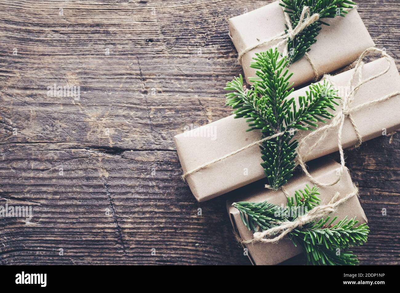 Moderne weihnachtliche Geschenke mit natürlichem Dekor auf altem Holzhintergrund. Drei Geschenkschachteln aus Bastelpapier mit Garn und grünen Zweigen. Saisonal Stockfoto