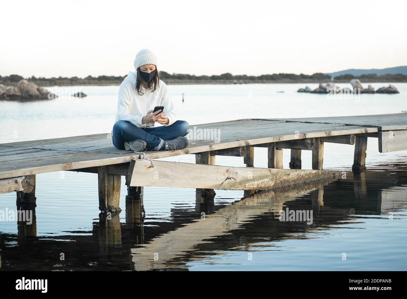 Einsame Frau lebt Natur sitzt auf einem Pier bei Sonnenuntergang Mit Smartphone zu chatten trägt Coronavirus sicheren Schutz medizinische Maske - Neue communicatio Stockfoto