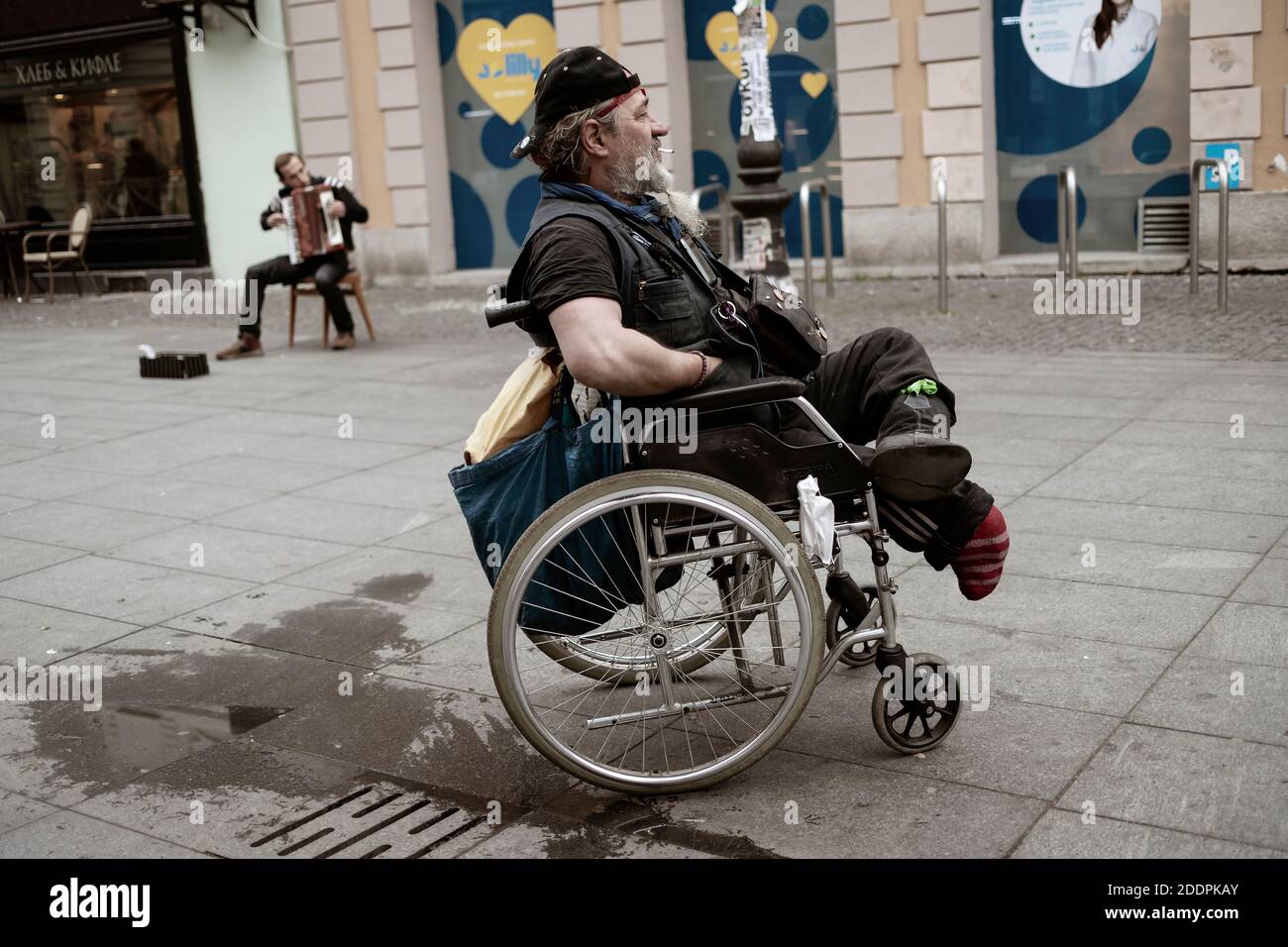 Belgrad, Serbien, 24. Nov 2020: Porträt eines Mannes mit Zigarette im Rollstuhl, der einem Straßenmusiker zuhört, der Akkordeon spielt Stockfoto