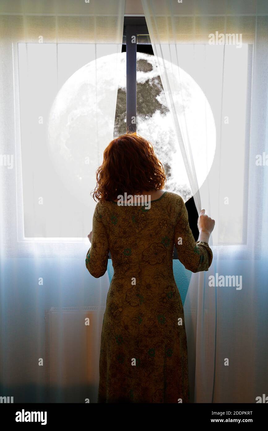 Frau, die auf den Supermond zwischen den Fenstervorhängen schaut Stockfoto