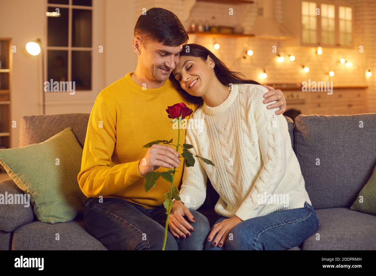 Junge lächelnde Mann Freund geben rote Rose als Symbol für Liebe zu seiner hübschen Freundin auf dem Sofa zu Hause Stockfoto