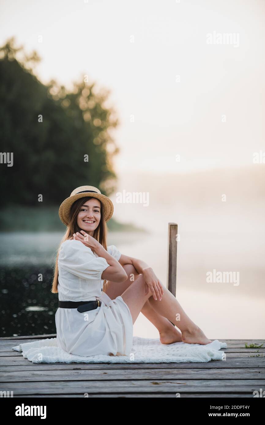 Attraktive Frau trägt weißes Kleid und Hut in der Nähe des Sees sitzen. Warme und gemütliche Sommermorgendatmosphäre. Stockfoto